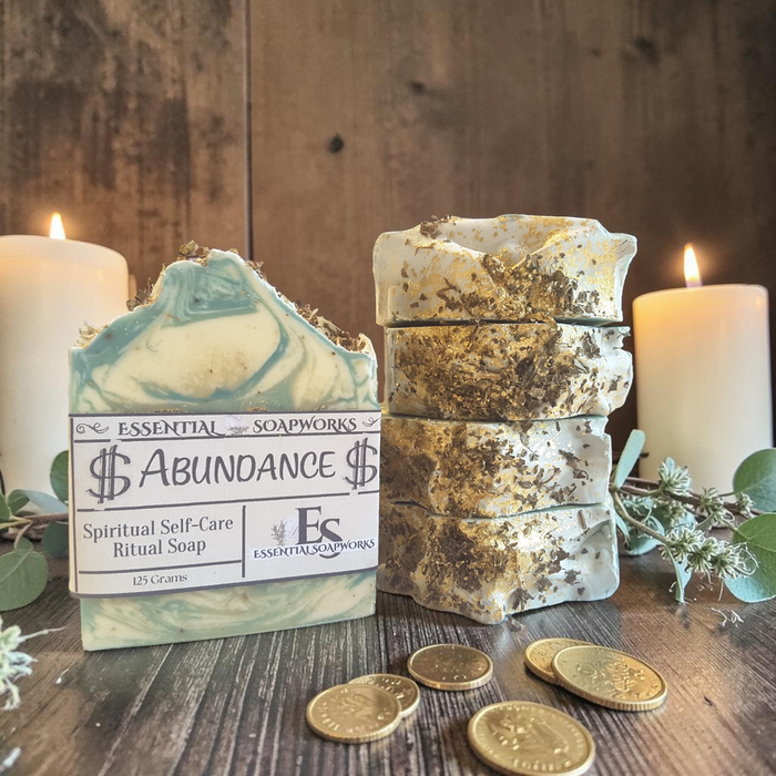 Two bars of Essential Soapworks 'Abundance' spiritual self-care ritual soap displayed on a rustic wooden surface with burning candles, eucalyptus sprigs, and gold coins. One bar is shown upright with its paper label visible, and the other is stacked in four pieces to show texture.