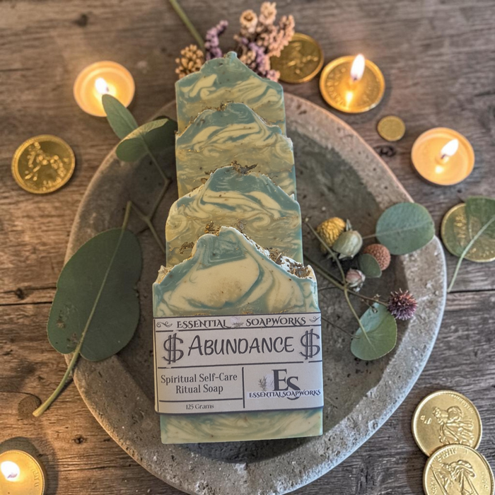 A bar of artisanal soap labeled '$ Abundance $' by Essential Soapworks, styled on a round stone tray with lit candles, gold coins, and eucalyptus leaves on a rustic wooden table.