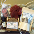 Autumn self-care ritual kit containing multiple wellness and spa items: a bottle of Autumn Air ritual oil, a bar of Essential Soapworks pumpkin spice soap, a pouch of Turmeric Tea Blend, a mesh red loofah, a candle in a white bag, and a small match pack, arranged in brown paper filler.
