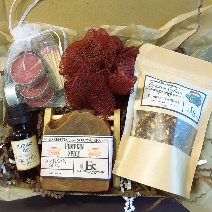 Autumn self-care ritual kit containing multiple wellness and spa items: a bottle of Autumn Air ritual oil, a bar of Essential Soapworks pumpkin spice soap, a pouch of Turmeric Tea Blend, a mesh red loofah, a candle in a white bag, and a small match pack, arranged in brown paper filler.