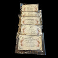 Five clear plastic bags of witchy Herbs for Banishing spells and rituals arranged in a vertical stack on a black background. Each bag has a cream-colored label with ornate floral borders and text including Angelica Root, Hyssop, Wormwood, Rue, and Mandrake Root.