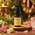 A 10 ml amber glass bottle of Beltane 100% Pure Essential Oil Blend with a black cap and yellow label, displayed on a rustic wood slice surrounded by pink and white flowers, green leaves, a lit candle, and a crystal cluster.