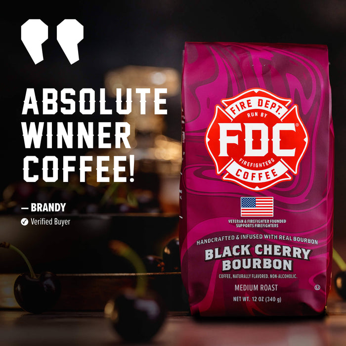 A bag of FDC Fire Department Coffee Black Cherry Bourbon medium roast coffee stands on a dark table with cherries in the foreground. The packaging is a vivid magenta-pink with a red and white Fire Department shield logo.