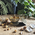 Decorative bronze incense burner with a smoking cone on a wooden surface with plants and crystals.