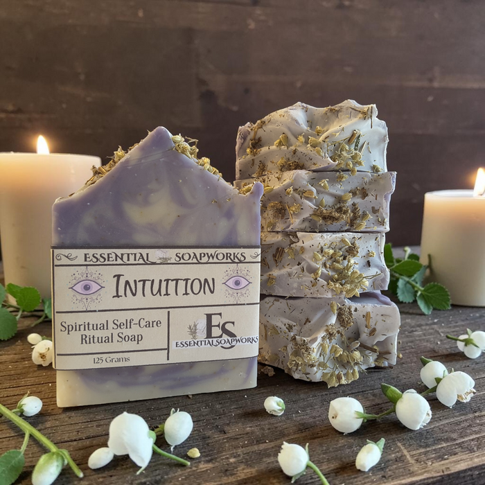 A bar of lavender and white marbled soap labeled 'Intuition' by Essential Soapworks, styled on a rustic wooden surface with three stacked bars, lit candles, and white blossoms.
