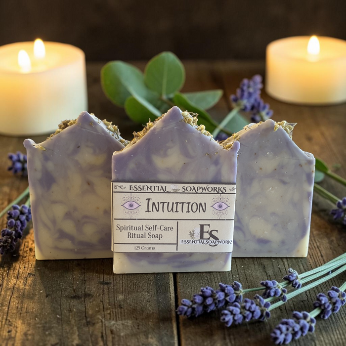 Three lavender and white marbled ritual soaps from Essential Soapworks are arranged on a rustic wooden surface with lit candles, green leaves, and purple lavender sprigs in the background.