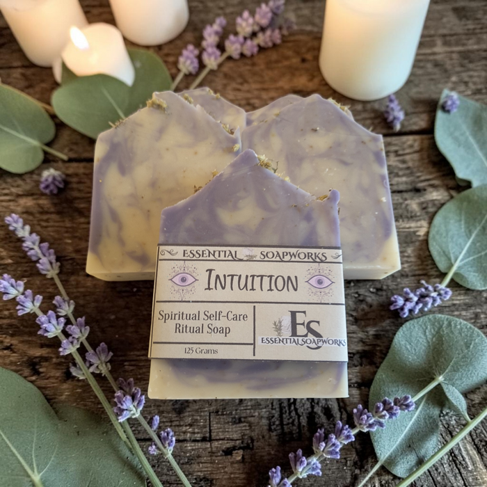 Three lavender and cream marbled ritual soaps arranged on a rustic wooden surface with white candles, eucalyptus leaves, and purple lavender sprigs. The front soap is labeled 'INTUITION' by 'ESSENTIAL SOAPWORKS'.