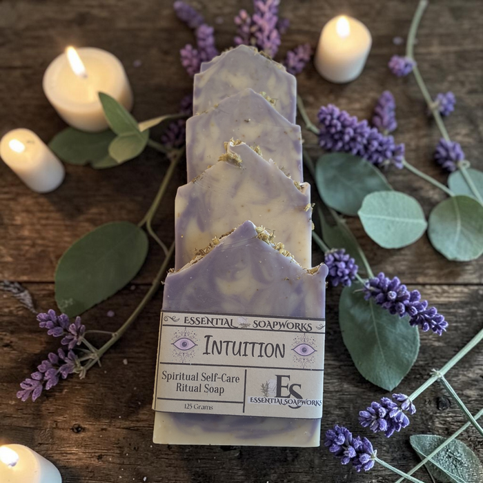 lavender and cream marbled ritual soap are stacked vertically on a rustic wooden surface, surrounded by lit white candles, green eucalyptus leaves, and purple lavender sprigs. The soap has a paper label that reads 'ESSENTIAL SOAPWORKS INTUITION'.