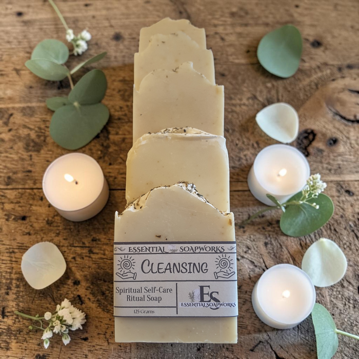 A stack of cream-colored ritual soap bars labeled 'Cleansing' by Essential Soapworks, arranged on a rustic wooden surface with lit tealights, eucalyptus leaves, and white petals.
