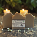 Three handmade rectangular soap bars with a central labeled bar reading ESSENTIAL SOAPWORKS Grounding, styled on a rustic wooden surface with lit candles and small white flowers in the background.