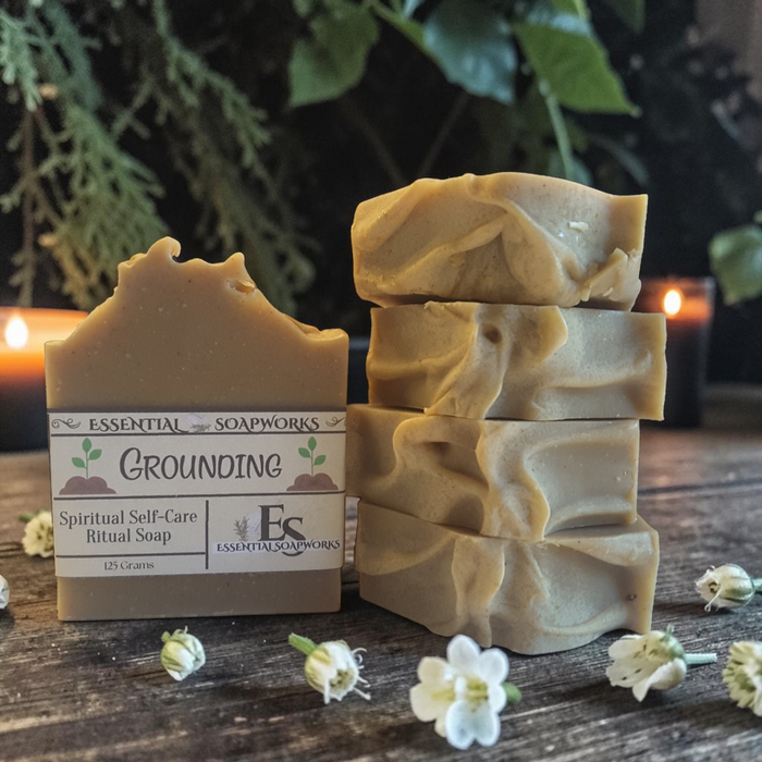 Four bars of handmade soap in a warm tan color, with one bar standing upright and three stacked. The upright bar has a label that reads 'ESSENTIAL SOAPWORKS GROUNDING Spiritual Self-Care Ritual Soap 125 Grams.