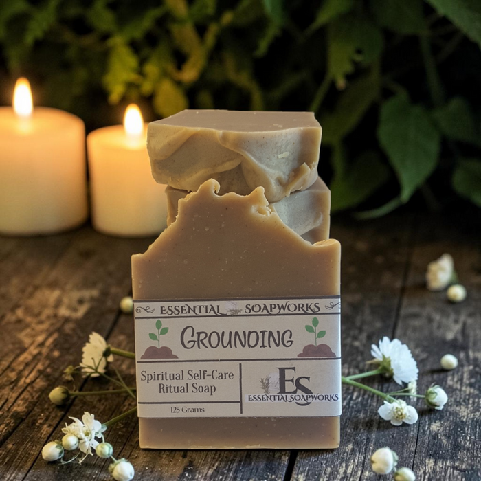 A handmade bar of soap labeled 'Grounding' by Essential Soapworks, styled on a rustic wooden surface with white flowers and glowing candles in the background.