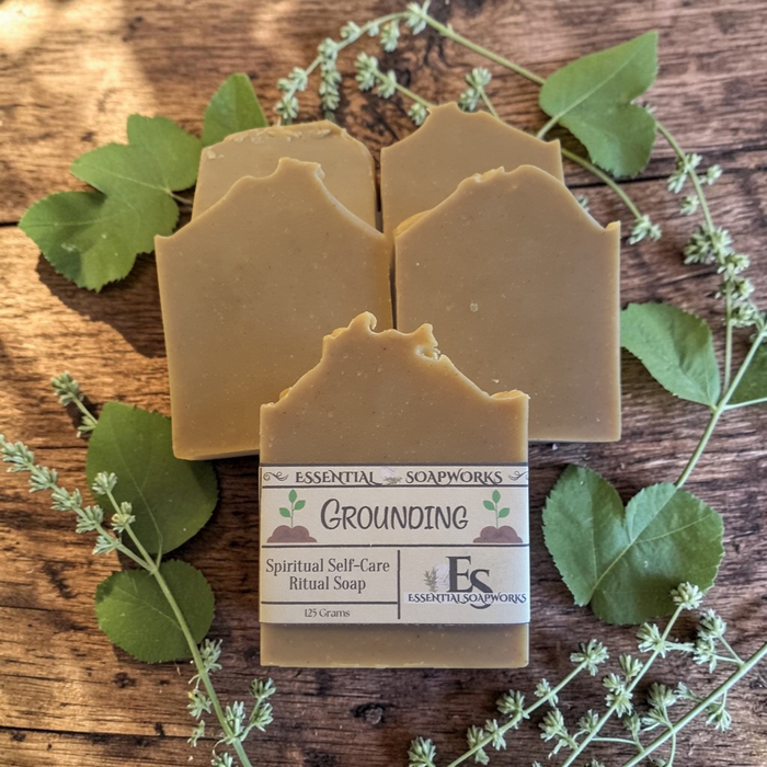 Four handmade soap bars arranged on a rustic wooden surface with green leaves and small flowering stems. The front bar has a paper label reading 'Essential Soapworks Grounding Spiritual Self-Care Ritual Soap'.
