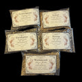 Five clear plastic bags of energy cleansing herbs for witchcraft with cream-colored labels are arranged on a solid black background. The labels feature botanical illustrations and names of the herbs: Lemongrass, Vetiver, Eucalyptus, Hyssop, and Wormwood.