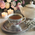 Evening wind down tea in a tea cup with floral design on a silver tray, teapot, candles, and flowers in the background.