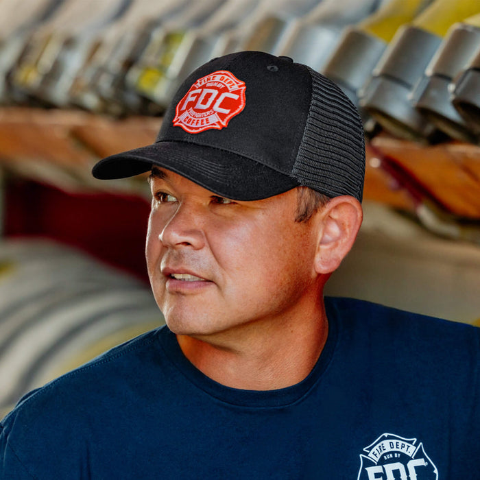 A man wearing a black mesh cap with the red Fire Department Coffee logo embroidery on the front and a blue shirt with white text, standing in front of a blurred background.
