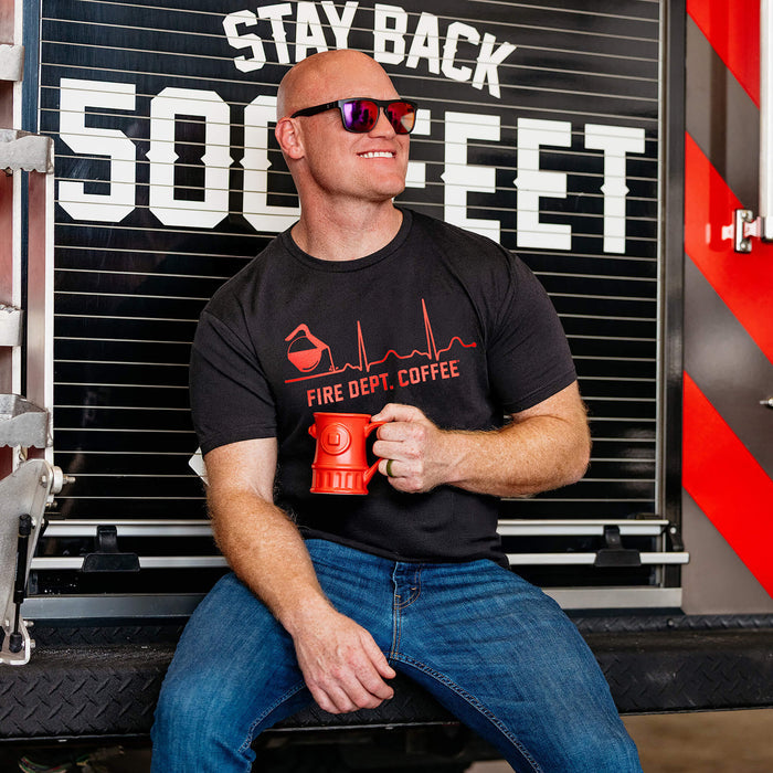 A man wearing a black Fire Dept. Coffee EKG T-shirt with a red design of a coffee pot pouring into a heartbeat line and “Fire Dept. Coffee” text underneath, holding a red hydrant mug while seated on the back of a fire truck.