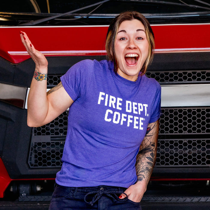 A woman wearing a purple t-shirt with white 'FIRE DEPT. COFFEE' text in front of a red and black fire truck.