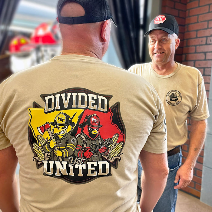Two men wearing tan graphic t-shirts in a casual indoor setting. The man in the foreground shows the back of his shirt with a large graphic reading 'DIVIDED AND UNITED' and two firefighter characters.