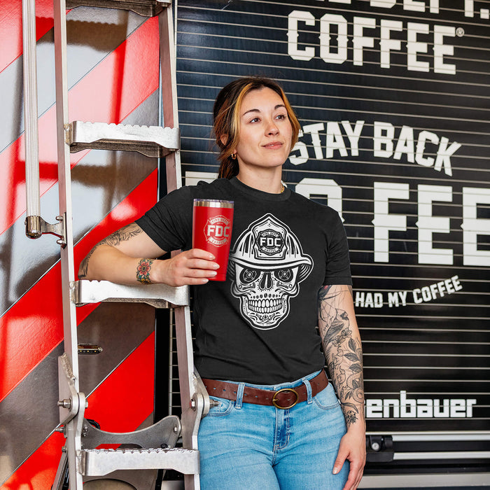 Woman wearing a black t-shirt with a skull design wearing a firefighter helmet, holding a red cup, and leaning on the back of a fire truck.