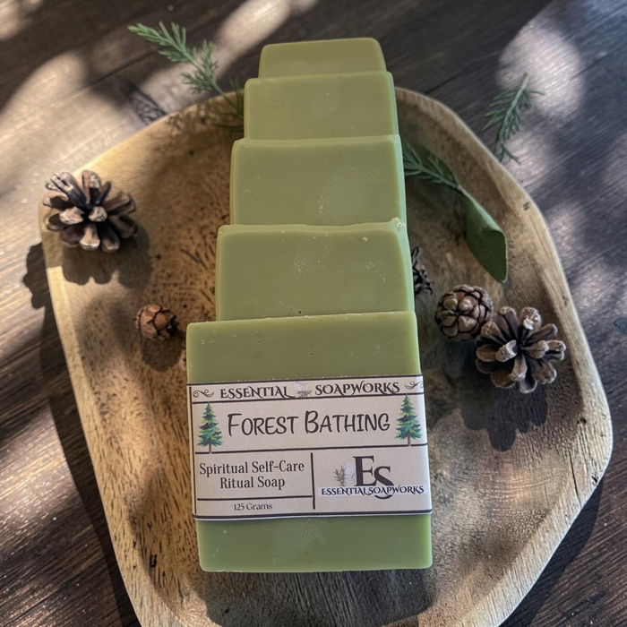 Five bars of Essential Soapworks Forest Bathing ritual soap are stacked on a rustic wooden tray, surrounded by pinecones and evergreen branches on a dark wood surface.