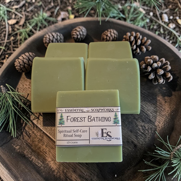 Four bars of olive green ritual soap arranged on a rustic wooden tray with pinecones and evergreen branches. The front bar is wrapped in a paper label that reads 'Essential Soapworks Forest Bathing Spiritual Self-Care Ritual Soap'.