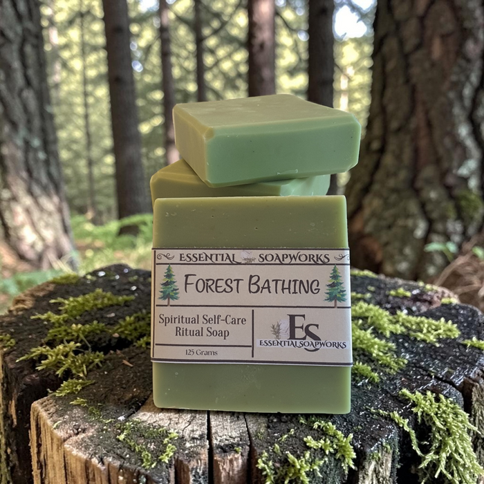 Three bars of green ritual soap stacked on a mossy tree stump in a forest setting. The front bar has a white paper label that reads 'ESSENTIAL SOAPWORKS Forest Bathing Spiritual Self-Care Ritual Soap'.