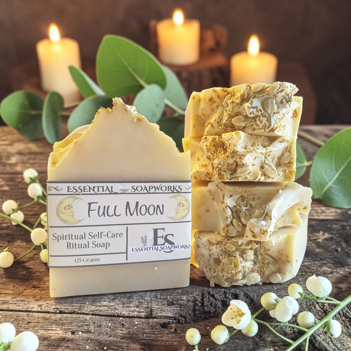 A bar of Essential Soapworks Full Moon Spiritual Self-Care Ritual Soap stands next to three stacked cut bars of the same handmade soap on a rustic wooden surface, with green leaves and white flowers around it and lit candles blurred in the background.
