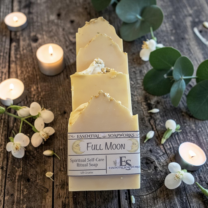 Four bars of Essential Soapworks Full Moon Ritual Soap arranged upright on a rustic wooden surface, surrounded by lit tea lights, white flowers, and green eucalyptus leaves.