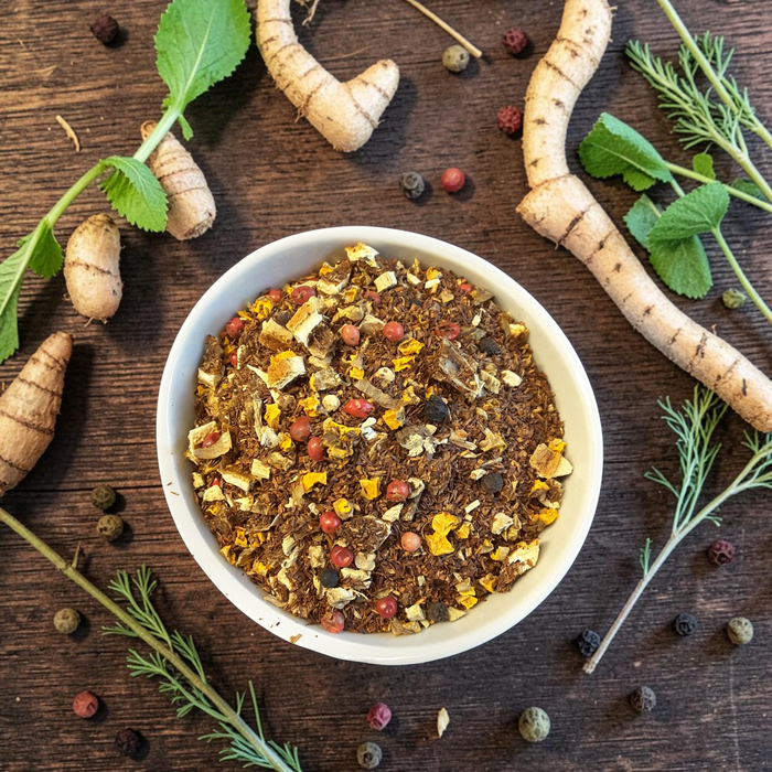 loose leaf turmeric tea in a bowl on a wooden surface with herbs, peppercorns and turmeric root