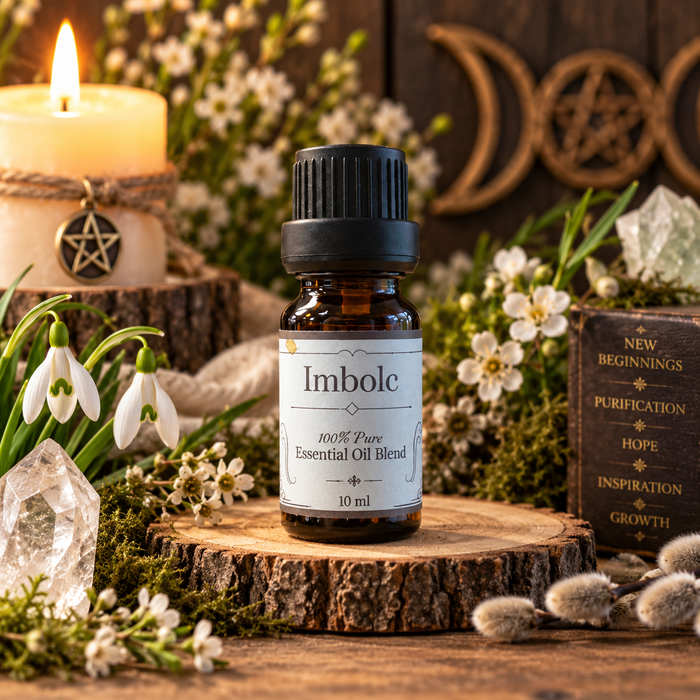A 10 ml amber glass bottle of Imbolc essential oil blend with a black cap and white label, displayed on a rustic wood slice. The background is a styled scene with a burning candle, snowdrop flowers, crystals, and a wooden book.