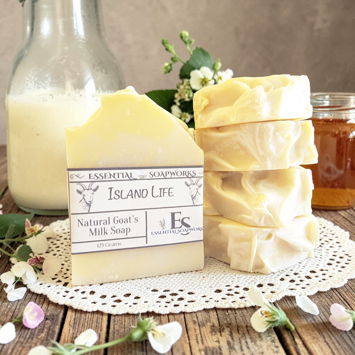 Goat milk soap bars on a wooden surface with flowers and a bottle in the background.