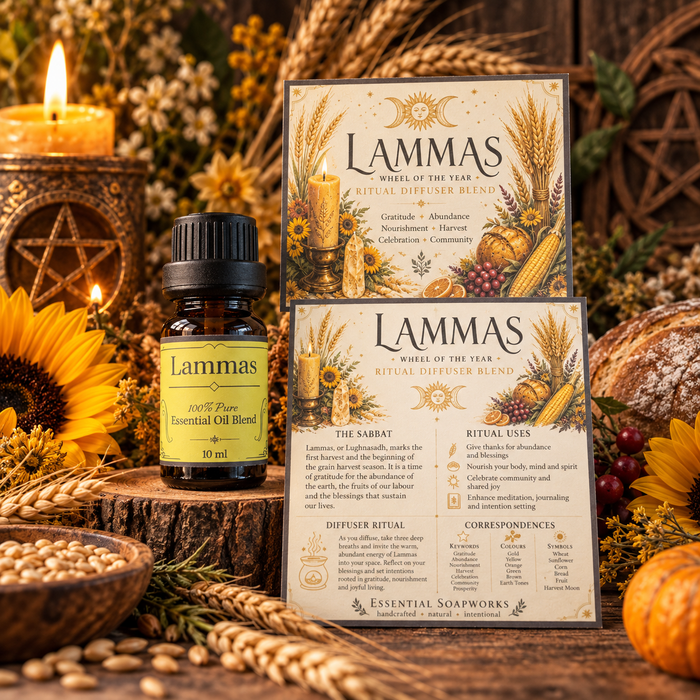A small amber glass essential oil bottle labeled Lammas Wheel of the Year Ritual Diffuser Blend stands beside two matching printed cards with illustrated packaging, arranged on a rustic wood stump with candles, sunflowers, grains, and autumn decor in the background.