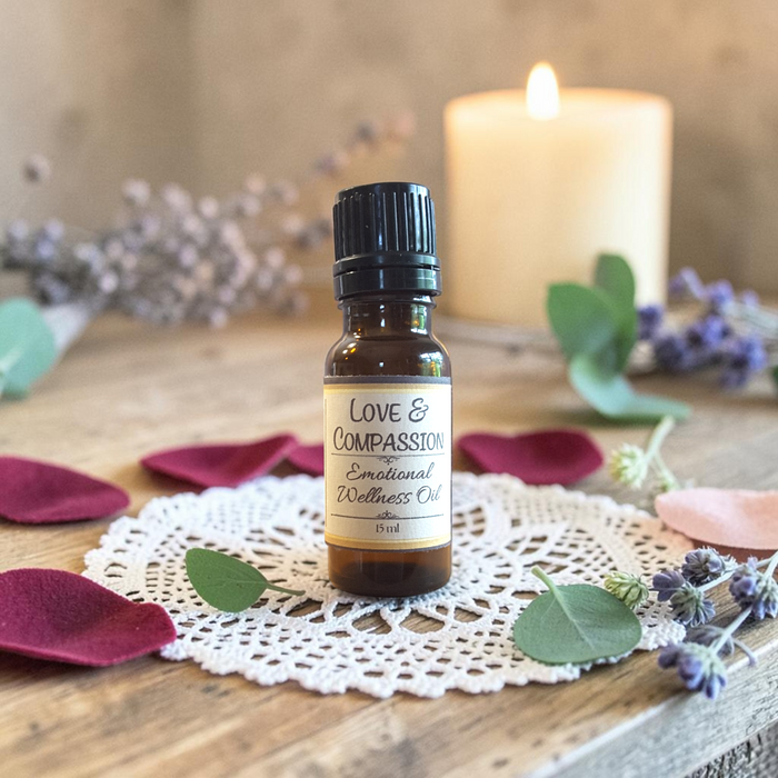 Love & Compassion Emotional Wellness Oil on a doily on a wooden surface with rose petals, greenery and a candle in the background