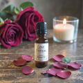 A small amber glass bottle labeled 'Love Potion' with a black cap stands on a rustic wooden surface, surrounded by pink rose petals. In the background, a lit white candle in a clear glass holder and dark red roses are visible.