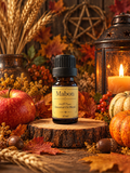 A 10 ml amber glass bottle of Mabon 100% Pure Essential Oil Blend with a black cap and cream label, displayed on a rustic wood slice. The bottle is surrounded by autumn props including a red apple, pumpkins, acorns, dried wheat, and a glowing candle in a lantern.