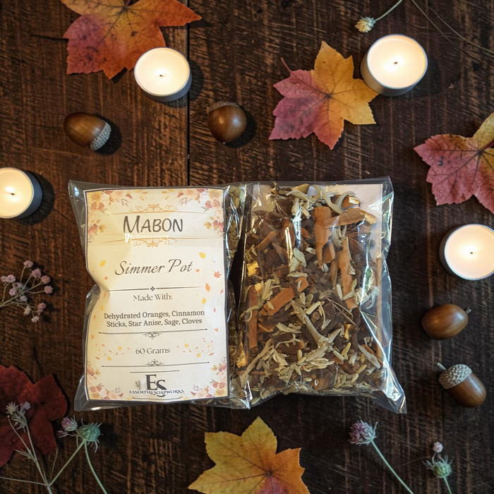 Two bags of Mabon simmer pot herbs arranged on a dark wooden surface with scattered autumn leaves, acorns, and small lit candles. The left bag has a cream label reading 'MABON Simmer Pot' and the right bag contains dried herbs and spices