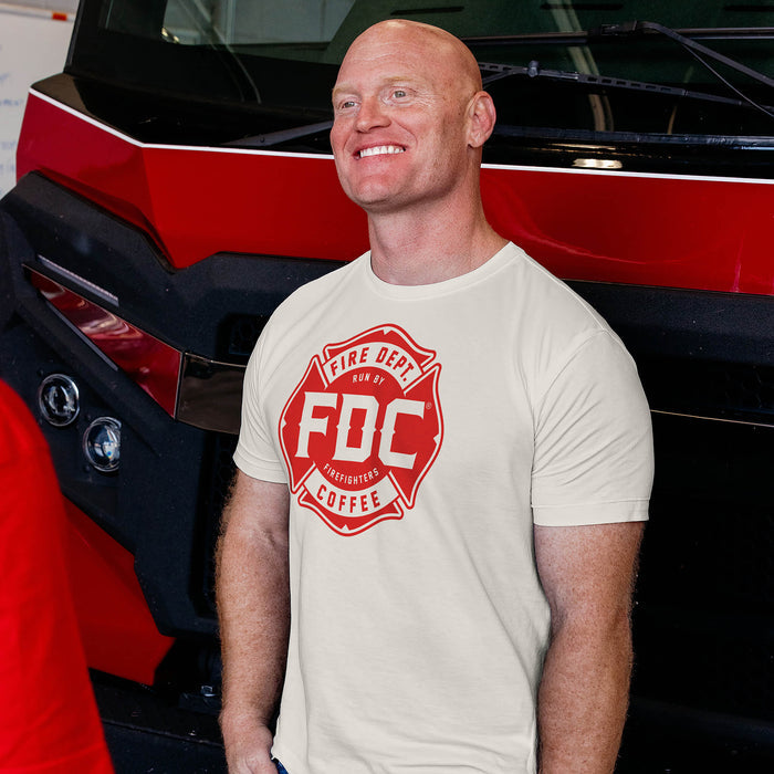 A man wearing a vintage white Fire Dept. Coffee T-shirt featuring a large red FDC Maltese cross logo, standing beside a red and black fire truck indoors.