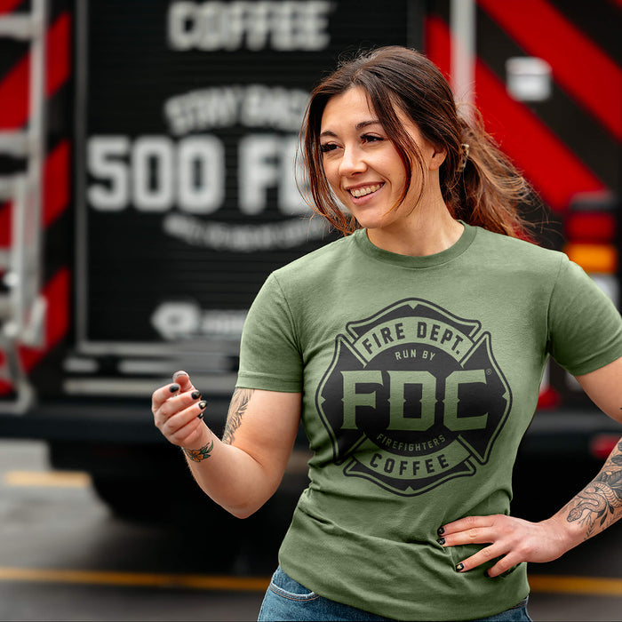 A woman wearing a military green Fire Dept. Coffee T-shirt featuring a large black Maltese cross logo, standing outdoors and a blurred fire truck is in the background