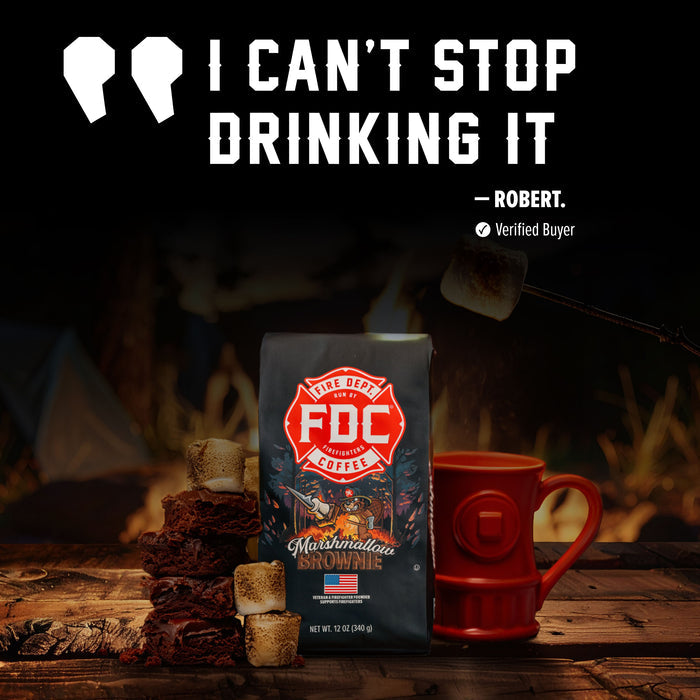 A bag of FDC Fire Department Marshmallows coffee sits on a rustic wooden table beside a red mug and marshmallows, with a campfire in the background and a large quote at the top reading 'I CAN'T STOP DRINKING IT - ROBERT. Verified Buyer'.
