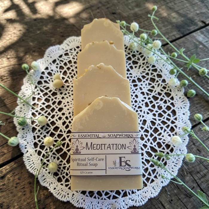 Four bars of meditation ritual soap standing upright on a white lace doily, surrounded by small white flowers on a rustic wooden surface. The front slice has a label reading 'Essential Soapworks' and 'Meditation'.