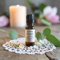 Mindful Presence Emotional Wellness Oil on a doily on a wooden surface with greenery and pink flowers and a candle in the background