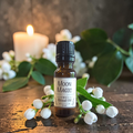 An amber glass bottle of Moon Magic Ritual Oil with a black cap and white label, styled on a rustic wooden surface with white flowers, green leaves, and a burning candle in the background.
