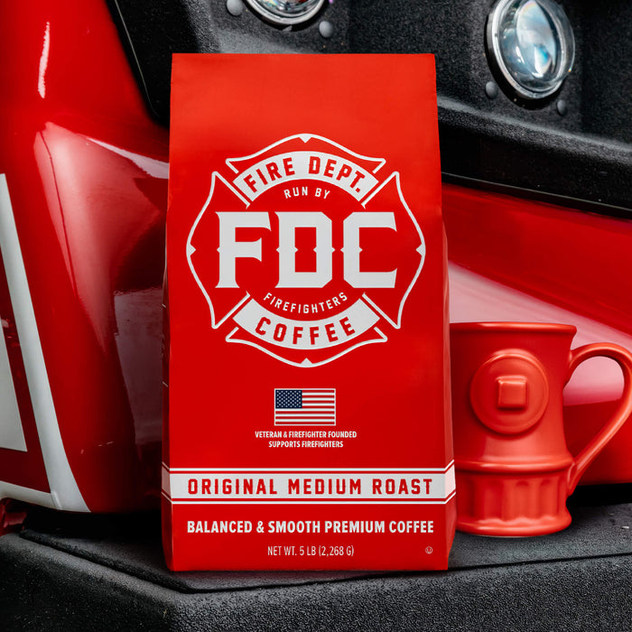 A red 5-lb coffee bag of Fire Department Coffee's Medium Roast Coffee stands on a black surface with a red fire hydrant-shaped mug beside it, set against a red and black fire truck background.