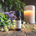 A small amber glass bottle labeled 'PEACE & CALM Ritual Oil 15 ml' with a black cap, placed on a rustic wooden surface with white flowers, green leaves, and a lit candle in the background.