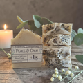 Essential Soapsworks Peace & Calm ritual soap shown as one cut bar beside a stack of four sliced bars, styled on a rustic wooden surface with a lit candle, greenery, and small white berries.