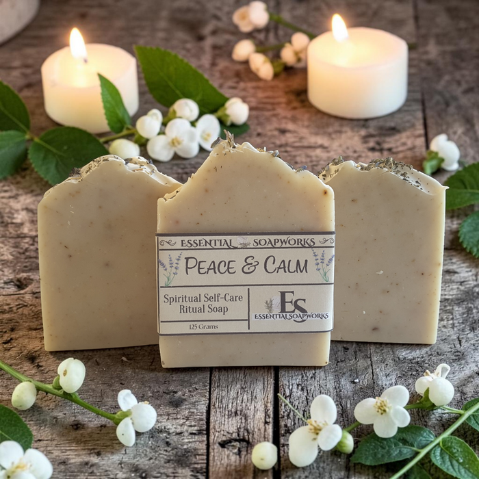Three bars of Essential Soapworks Peace & Calm ritual soap are arranged on a rustic wooden surface with lit white candles, green leaves, and small white flowers. The center bar is upright and shows the product label.