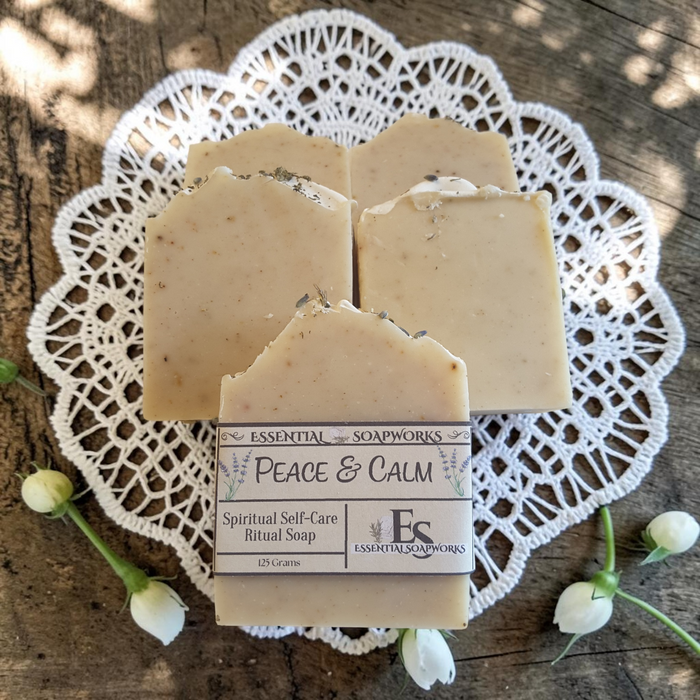 Four bars of Essential Soapworks Peace & Calm ritual soap arranged on a white lace doily over a rustic wooden surface, with small white flowers around the soap bars.