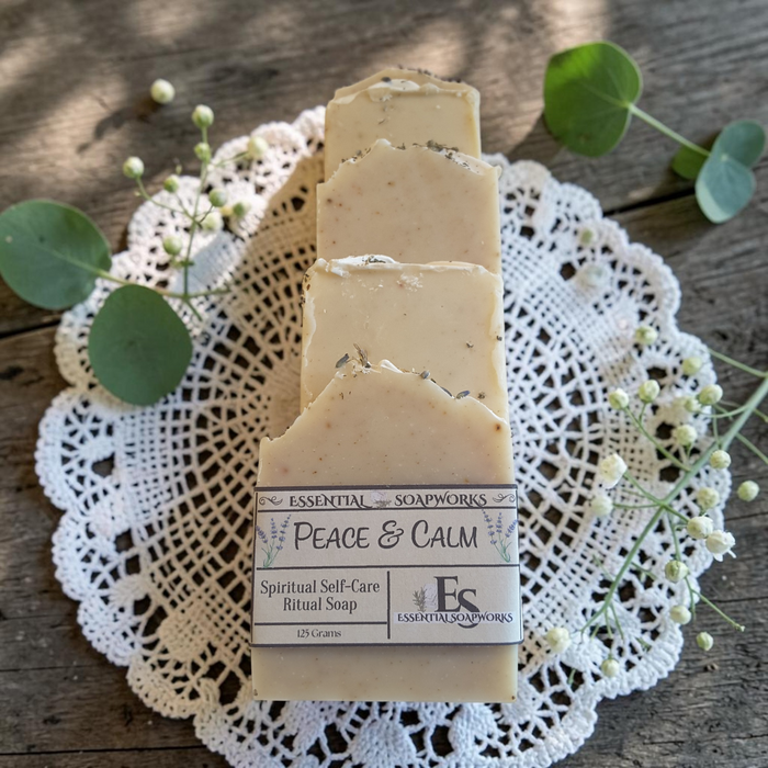 Three bars of Essential Soapworks Peace & Calm ritual soap displayed on a white lace doily over a rustic wooden surface, accented with green eucalyptus leaves and small white flowers.