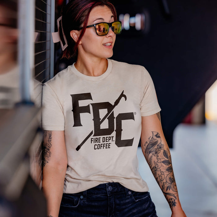 A woman wearing a vintage white Fire Dept. Coffee T-shirt featuring a large black FDC logo with a pike pole graphic running through it and small ‘Fire Dept. Coffee’ text below, leaning against a surface while wearing sunglasses and a blurred background.